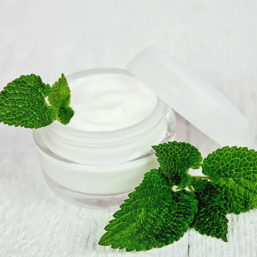 Open jar of white cream beside fresh lemon balm leaves on painted wood