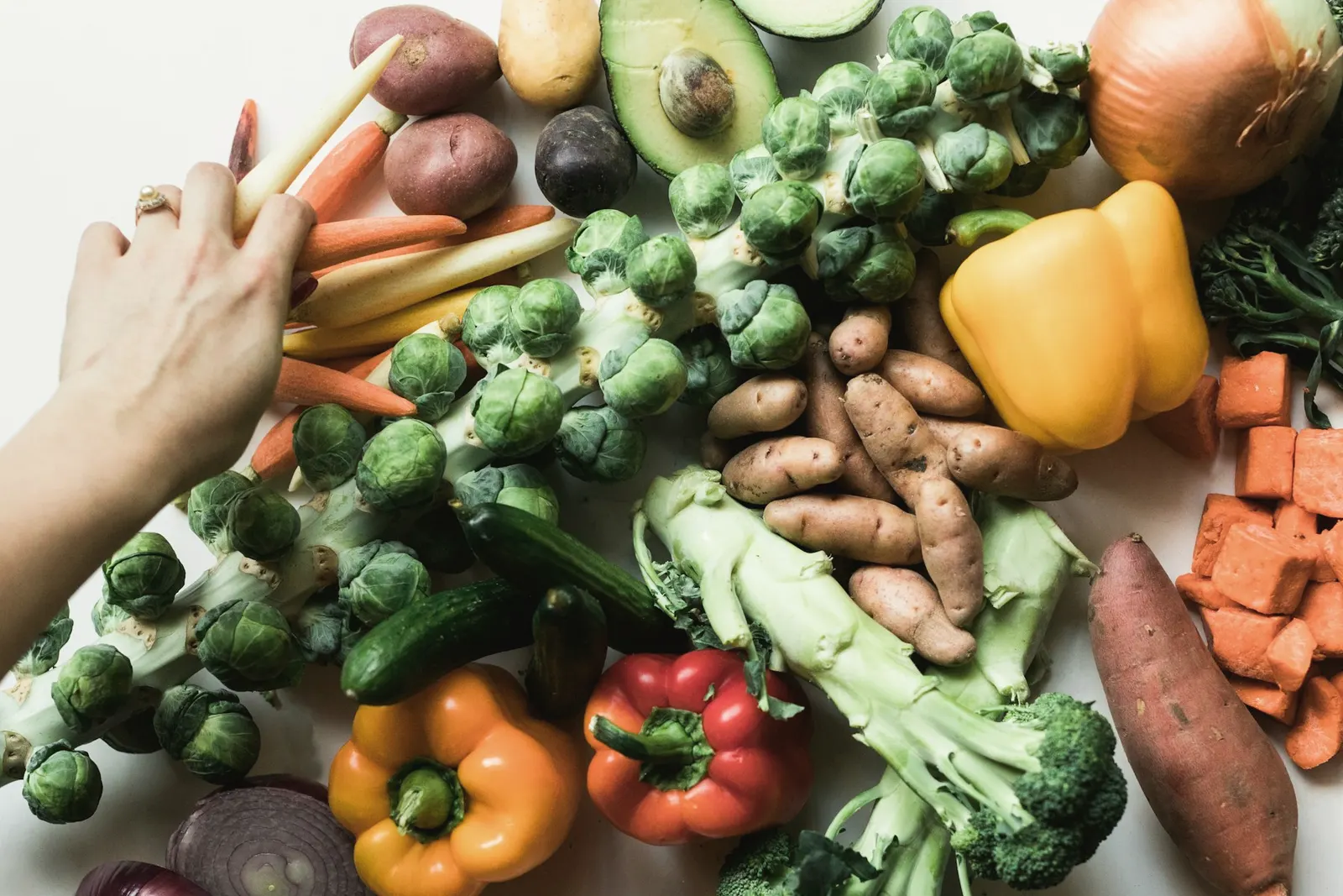 A hand reaching for fresh vegetables including sweet potato, capsicum, carrots and avocado
